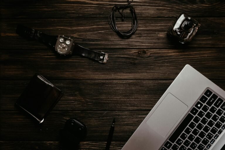 Flat lay of laptop, watch, wallet, and accessories on a dark wooden surface.