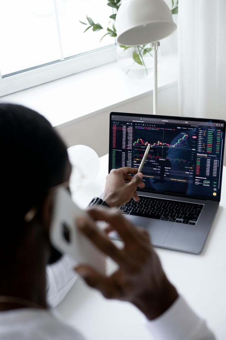 A businessman using a smartphone while analyzing stock market graphs on a laptop indoors.